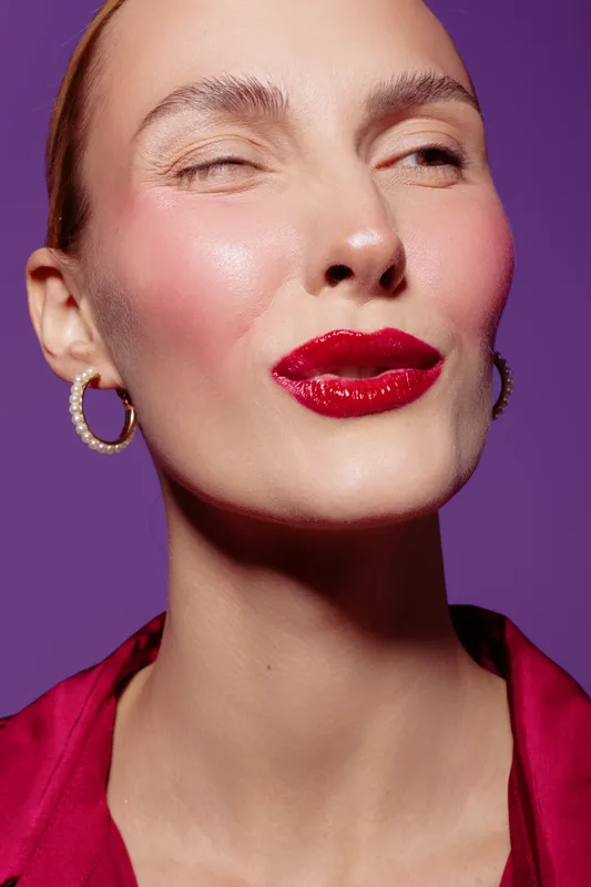 Close-up of a woman with glossy red lips and pearl earrings making a playful face against a purple background. Concept of beauty, makeup, fashion, and confident expression.