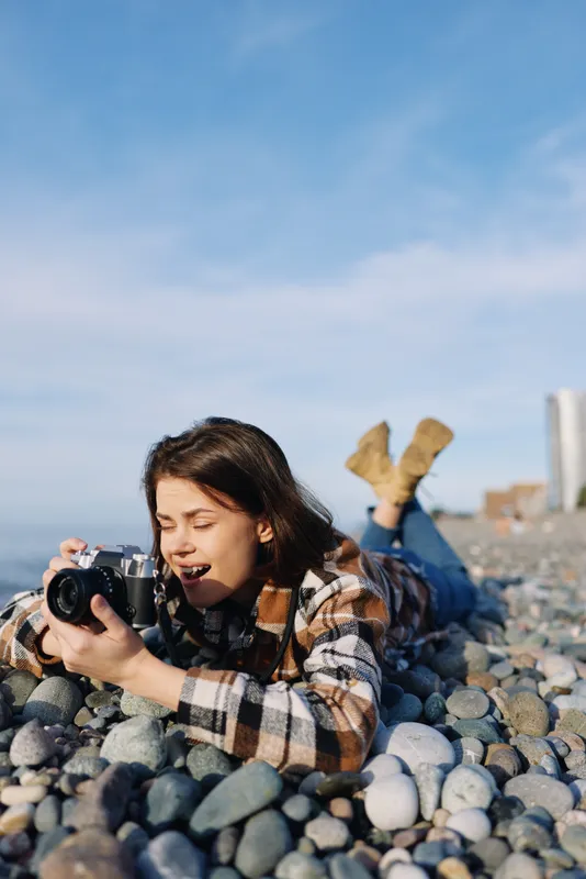 Woman photographer on beach lying among smooth stones with a camera, wearing a plaid shirt, capturing candid moments and coastal scenery under natural light