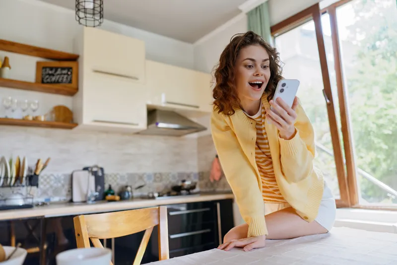 Cheerful woman in a bright kitchen wearing a casual yellow cardigan, laughing at her phone, warm sunlight across a cozy home, relaxed lifestyle moment, natural colors and candid mood