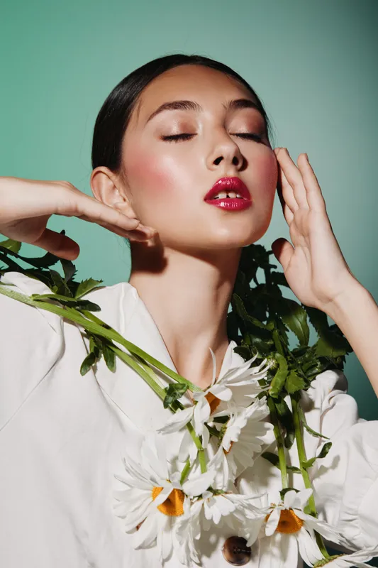 Young woman with glossy red lips poses elegantly with chrysanthemums on turquoise background. Fashion portrait showcases beauty, skincare, and creative floral styling influence.