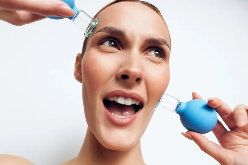 Young woman with clean skin applies serum and uses blue facial suction tool on her cheek, demonstrating skincare routine and beauty treatment on white background.