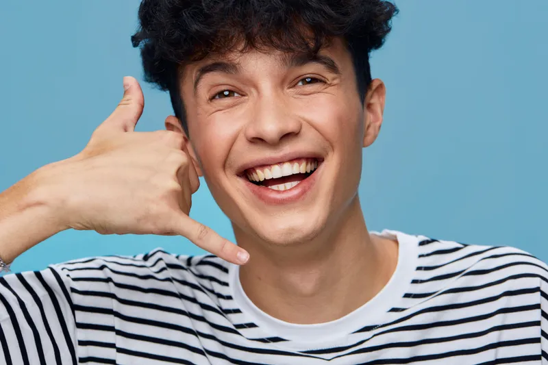 young man smiling shows call me gesture with hand near face against plain blue background, casual striped shirt, cheerful expression, close-up portrait
