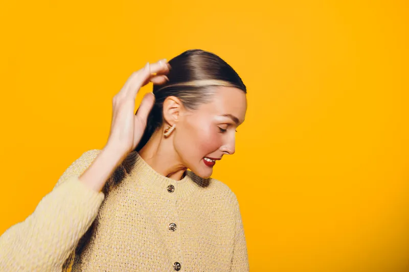 Young woman with slicked back hair wearing beige sweater looks down and touches head against bright yellow background. Concept of fashion, style, portrait, and modern beauty.
