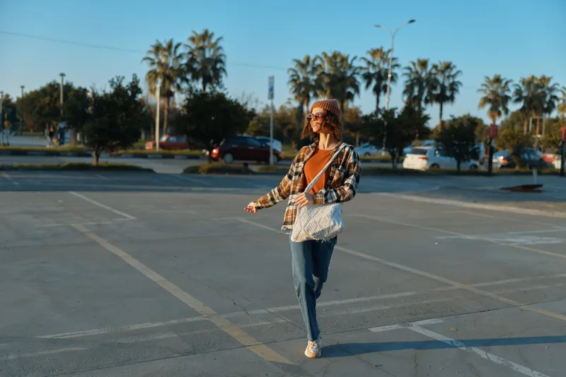 Woman walking in parking lot at golden hour, casual outfit with sunglasses and tote bag by palms; candid lifestyle portrait emphasizing authenticity, mindful living and emotional storytelling.