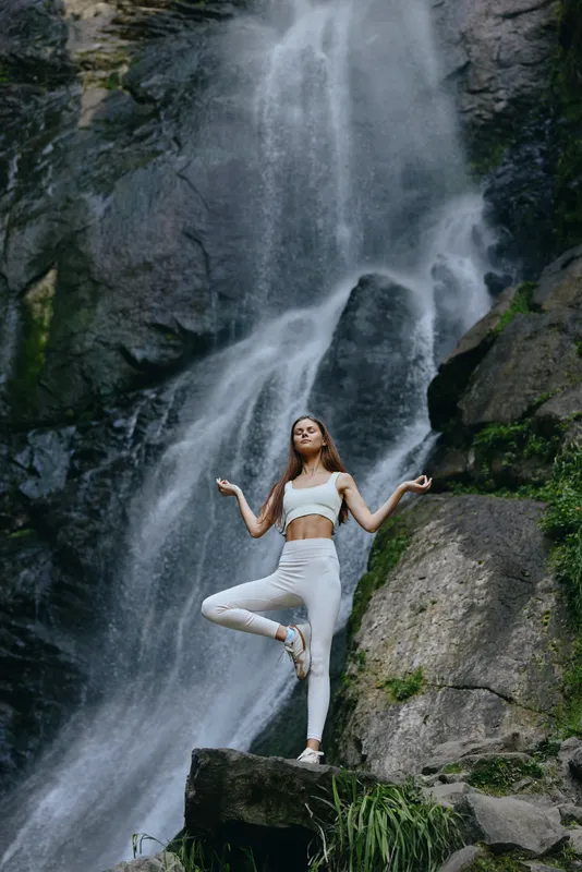 woman practicing yoga in nature with waterfall background, wearing white sportswear, demonstrating balance and calmness in a serene outdoor setting