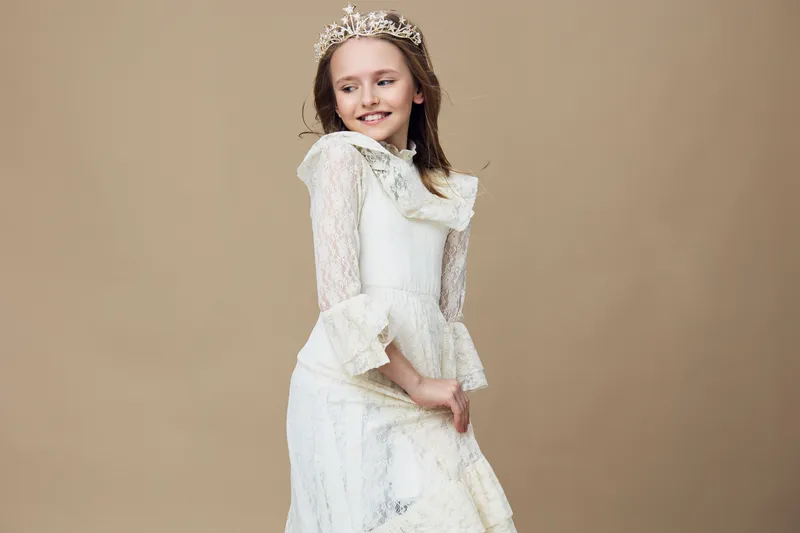 Young girl in a white dress and crown, smiling joyfully against a soft beige background, perfect for concepts of childhood and celebration