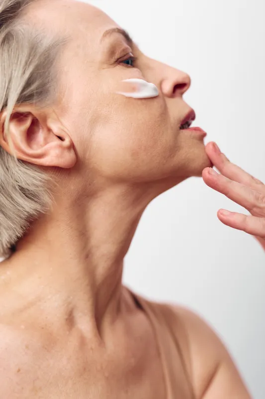 Mature woman with short hair applies face cream on cheek, showing skin care routine and natural beauty. Concept of aging gracefully, wellness, and self-care with soft lighting.