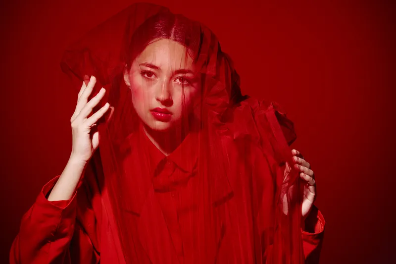 Portrait of a stylish young woman wrapped in layered red tulle fabric on a red background. High fashion avant-garde beauty concept with bold makeup and dramatic lighting.