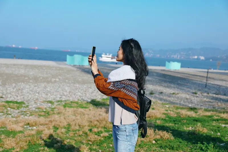 Woman photography smartphone beach outdoor ocean scene, casual scarf backpack, sky sunlight shore sand coast seascape view moment image capture inspiration leisure fashion lifestyle mood blue horizon
