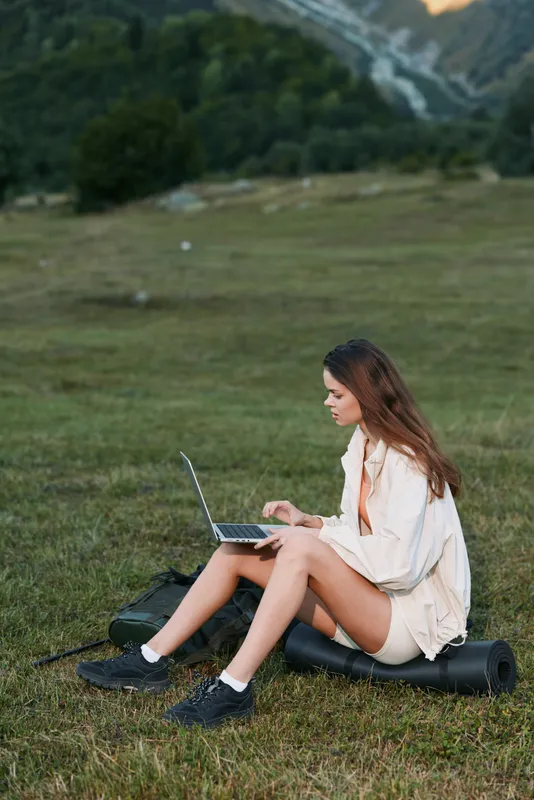 A cheerful young woman sits on grass with a laptop in a scenic outdoor setting. She focuses on the screen, exuding calm productivity and a relaxed, creative mood.