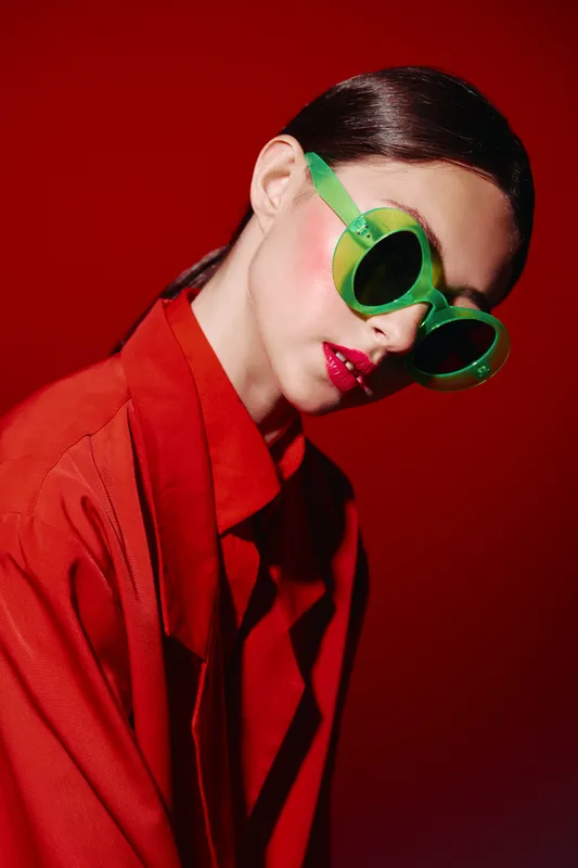 Fashion model with slicked hair and red makeup wearing green sunglasses and a red jacket poses against a red background. Concept for beauty creative fashion editorial.