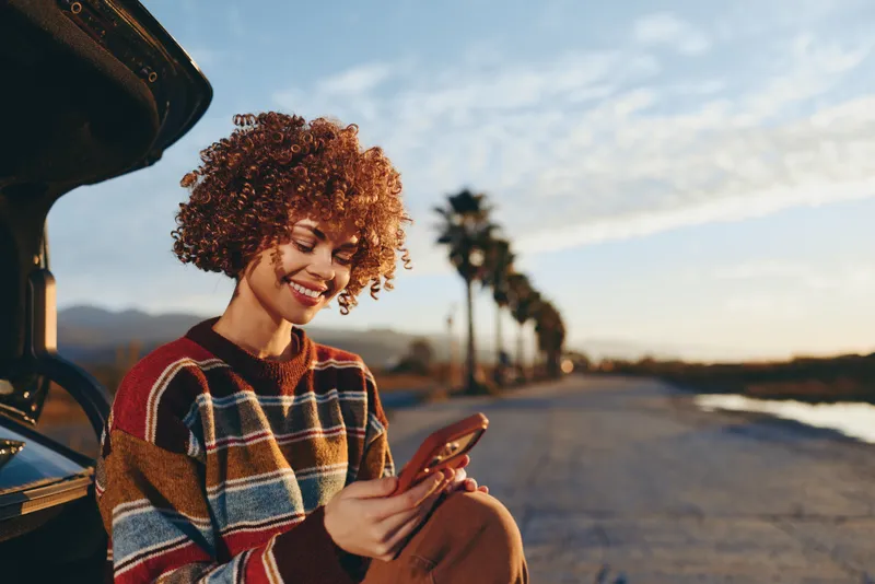 Woman lifestyle scene, smiling with curly hair, holding smartphone outdoors by street road at sunset, wearing plaid sweater, relaxed portrait, warm evening light, mobile communication