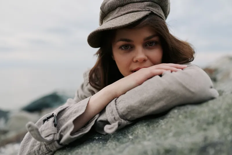 Young woman wearing a casual hat rests her arms on a rock outdoors. Soft natural light highlights her thoughtful expression in a relaxed, natural environment.
