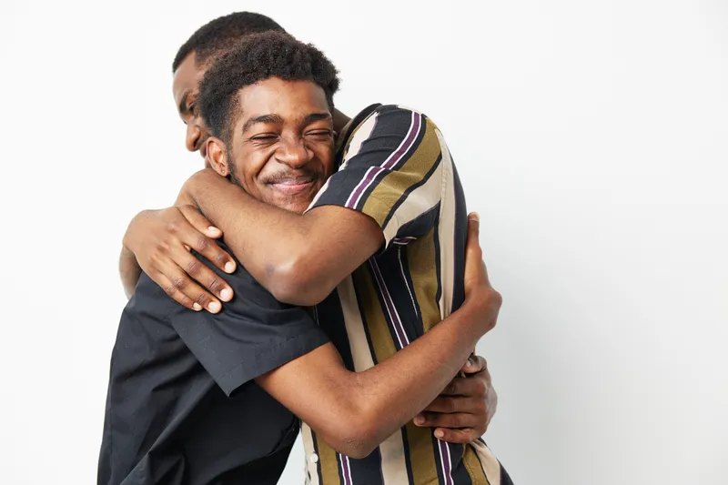 Two black men embrace warmly, displaying deep friendship and joy. Their smiles convey happiness and support in a light, neutral background.