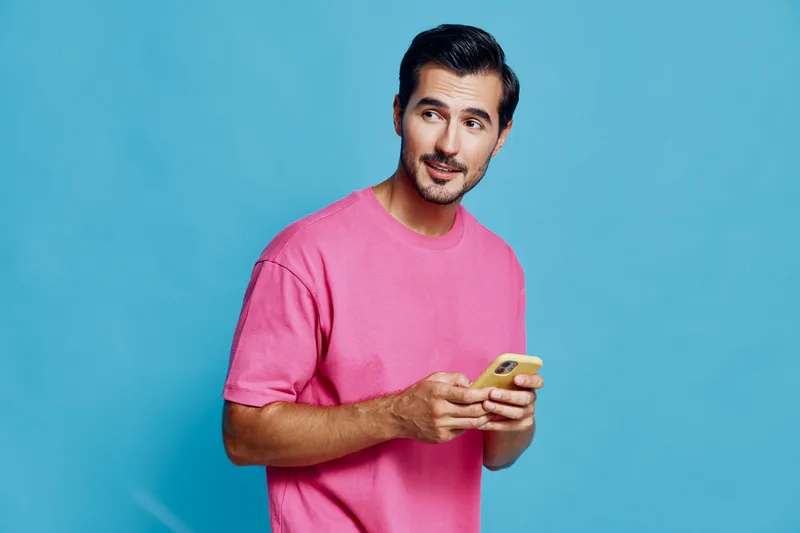 Full body smiling happy cheerful young man wear pink t-shirt look camera hold in hand use mobile smartphone isolated on vibrant blue background studio portrait. People lifestyle concept.