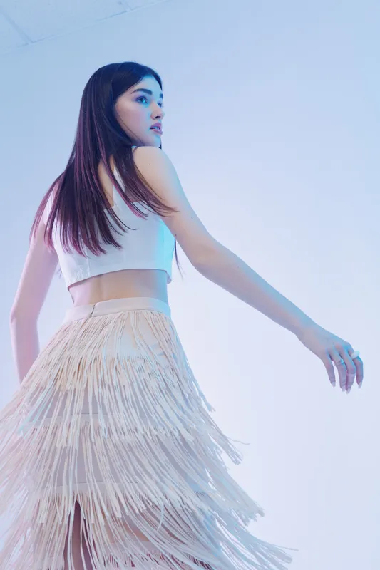Young woman in a white crop top and fringe skirt poses gracefully in a soft blue studio. Concept of beauty, fashion, elegance, and modern style with delicate lighting.