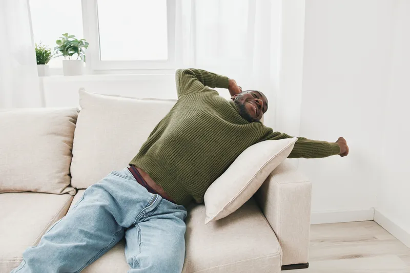 Relaxed Black man stretching on a sofa in a cozy living room, wearing a green sweater and light jeans Comfortable atmosphere promotes well being and relaxation