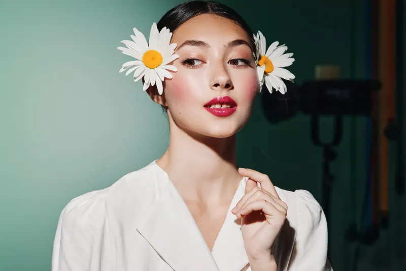 Beautiful young woman with glowing skin wearing white outfit and creative floral hair accessories, styled with vibrant makeup against a clean background, beauty fashion concept.