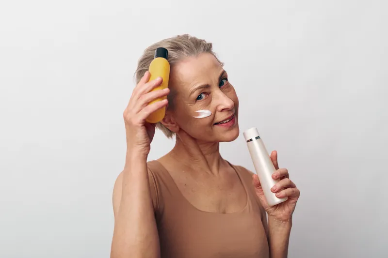 Smiling mature woman with natural skin applying cream on cheek holding two skincare bottles on light background. Concept of anti aging, beauty routine, healthy skin and self care.