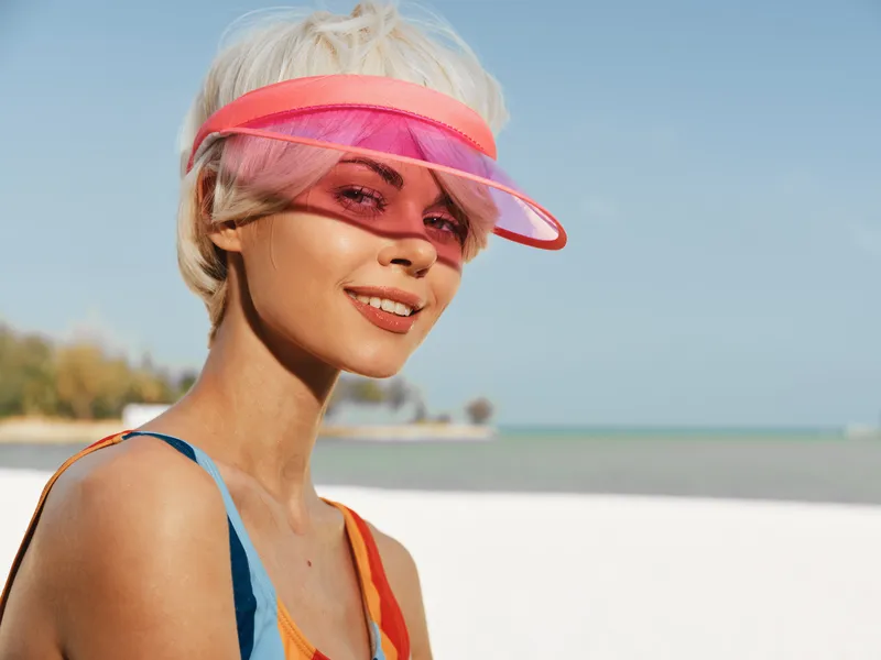 Portrait of a young woman smiling at the camera on a sunny beach, wearing a pink visor and colorful swimsuit, with clear blue sky and calm sea in the background.