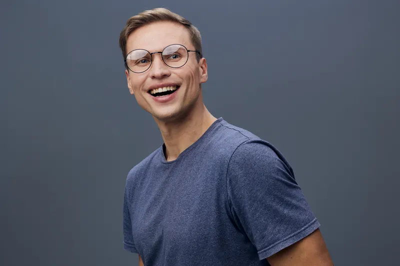 Smiling young man with glasses, wearing casual blue t-shirt, looking cheerful and confident, isolated on plain dark gray background, studio portrait. People lifestyle concept, positive emotion, casual