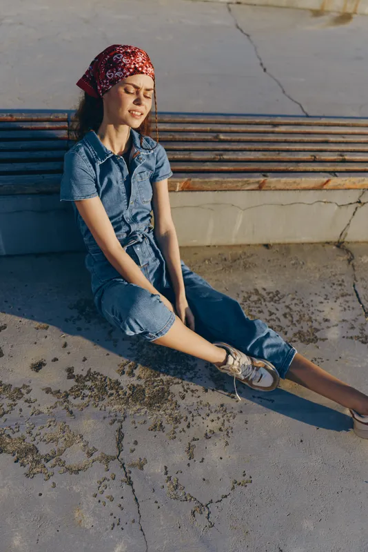 Lifestyle woman in modern boho-western denim street style sits relaxed on concrete ground in warm film color sunlight, wearing a red bandana and casual sneakers.