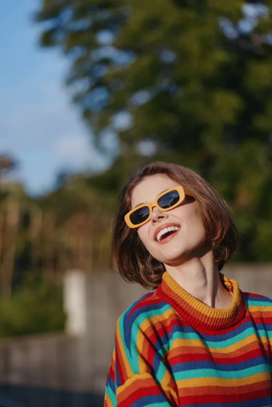 Woman smiling in striped sweater and yellow sunglasses outdoors, short hair portrait looking up with joyful expression, casual fashion knit, lifestyle travel vibe in green park.