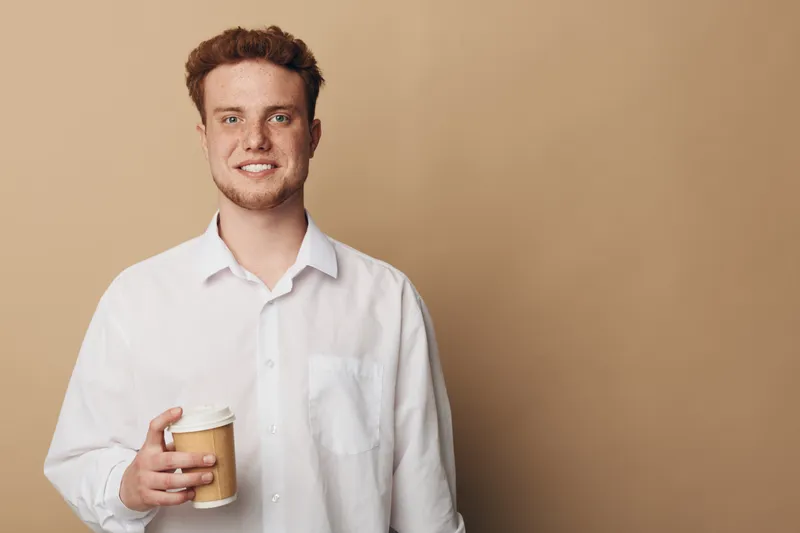 Smiling young man with red hair and beard holds disposable coffee cup, wearing white shirt against beige background. Concept of casual lifestyle, morning routine, and coffee break.