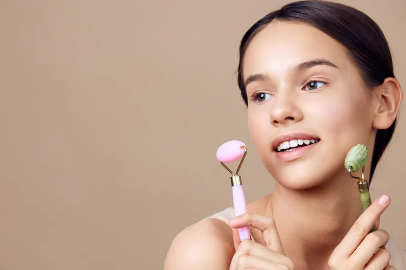 Young woman holding beauty rollers for skincare, promoting healthy skin and natural beauty with a cheerful expression against a beige background