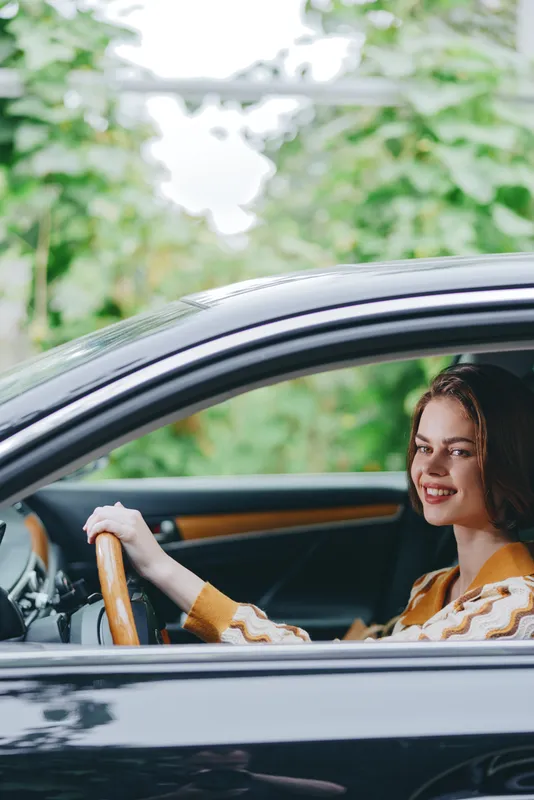A cheerful woman drives a car, hands on the steering wheel, wearing a cozy sweater, with bright daylight and lush green background.