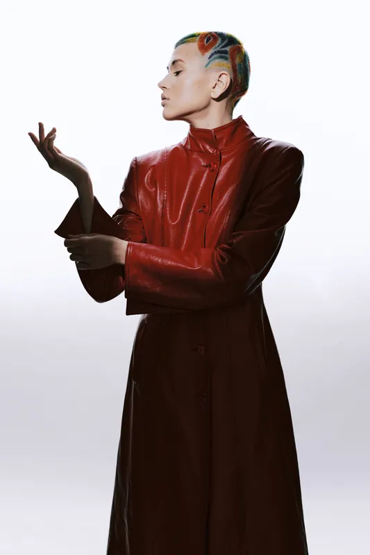 woman with colorful braided hair wearing a red leather coat poses elegantly against a bright white background with soft shadows and strong contrast