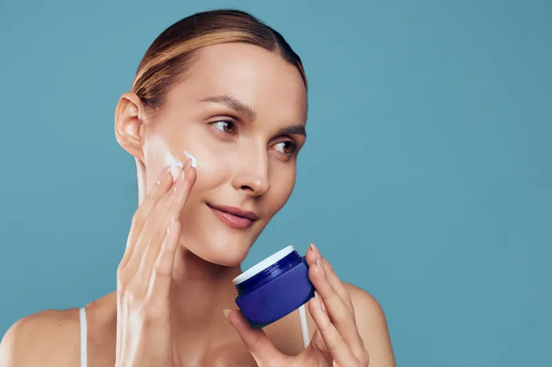 Young woman with clear skin applies moisturizer cream on her cheek against a plain blue background with a calm and focused expression.