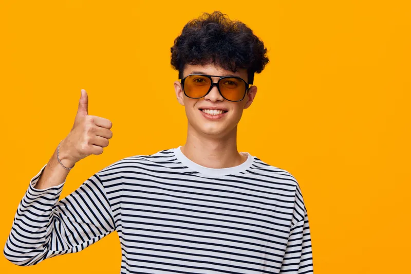 Smiling young man wearing orange sunglasses and striped shirt giving thumbs up with curly hair against bright orange background, casual style and positive mood.