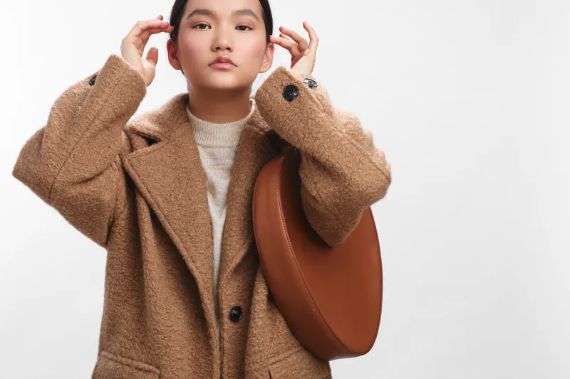 asian woman wearing a cozy beige coat stands against a plain backdrop, posing confidently as a fashion forward shopper, conveying warmth, style, and urban charm.