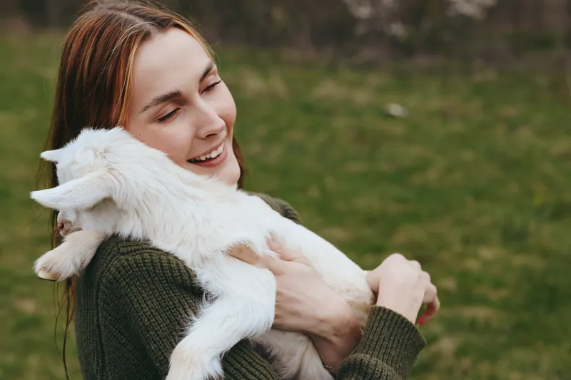 Lifestyle woman goat kid outdoors affection cuddle nature happiness photography. A candid scene of a smiling woman holding a goat kid in a grassy field, celebrating connection and gentle bonding.