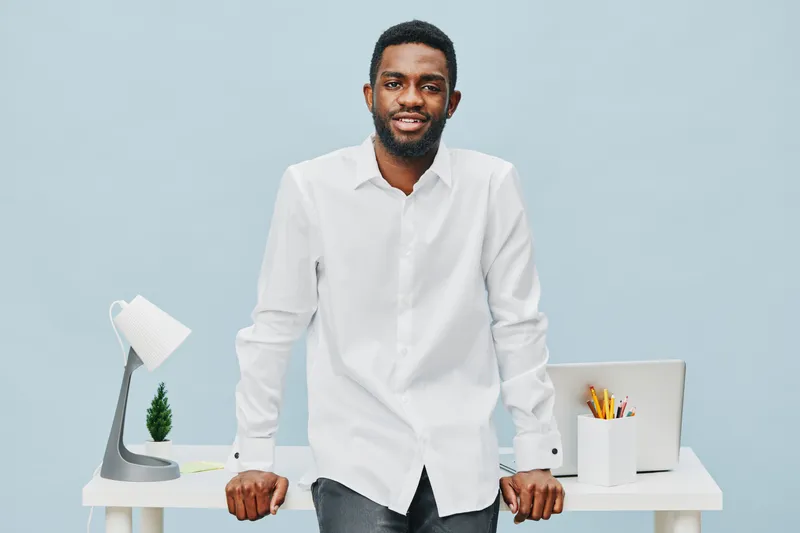 Confident young man with dark skin smiling in casual white shirt standing at modern office desk with laptop, stationery, and decorative plant isolated on light blue background. Business professional c