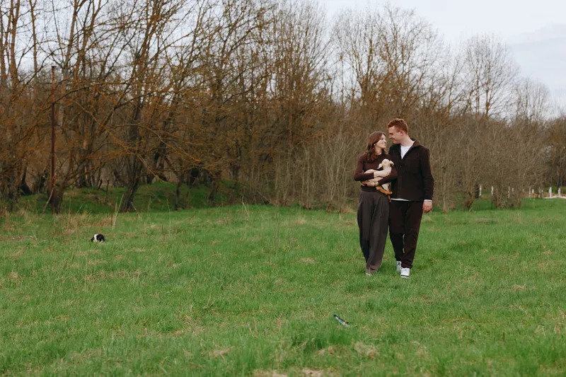 couple, outdoor, park, lifestyle, dog, companionship, stroll, nature: A relaxed scene shows a couple walking through a sunlit field, the woman cradling a small dog, capturing everyday affection and