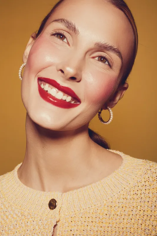 Smiling woman with red lipstick and hoop earrings wears yellow knitted sweater. Close-up portrait on warm yellow background. Concept of beauty, fashion, and positive emotions.