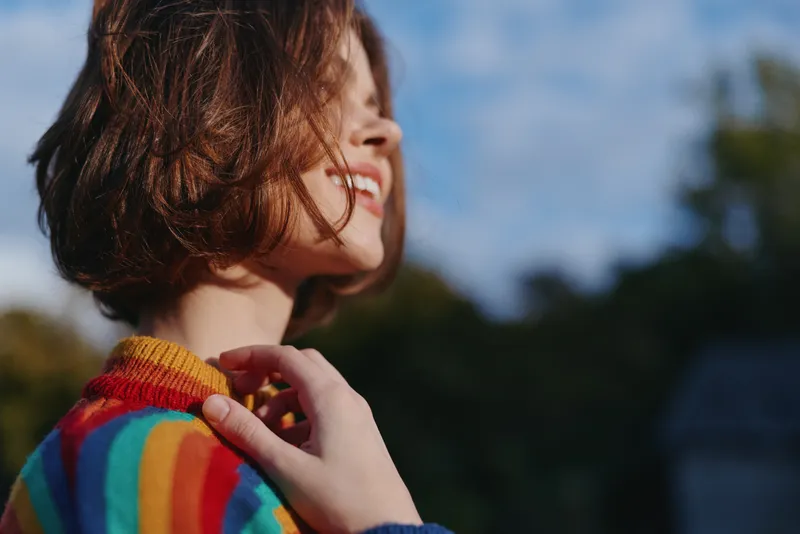 Woman smiling in a colorful striped sweater, short hair, young adult portrait outdoors in nature, casual outfit and relaxed pose, hand near collar, joyful lifestyle and travel vibe.