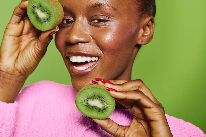 Smiling African woman with dark skin holds two halves of kiwi fruit near her face against green background. Concept of healthy lifestyle, natural beauty, fresh fruit, and wellness.