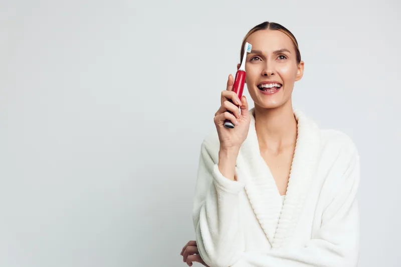 Smiling woman in white bathrobe holding electric toothbrush near face on light background. Concept of dental hygiene, morning routine, and self-care with bright soft lighting.
