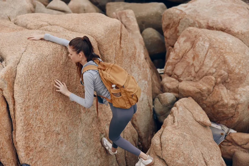 Young female climber in athletic wear navigating rocky terrain Her determination is evident as she climbs with a brown backpack on a sunny day