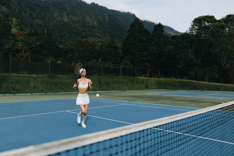 Young athletic woman enjoying tennis on a sunny court with mountains in the background, wearing a white skirt and top, displaying joy and focus