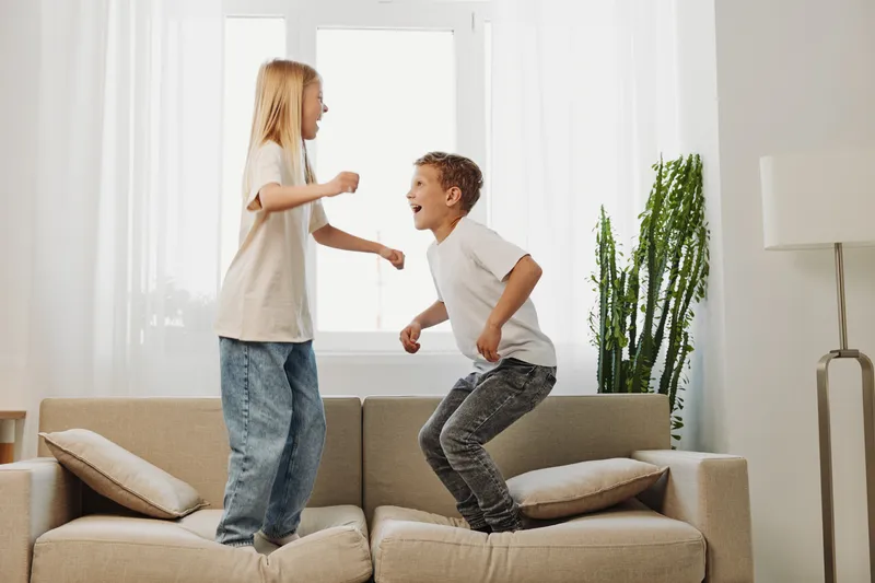 Happy children jump excitedly on a beige sofa in a bright living room. Siblings enjoy playful moments together, expressing joy and carefree childhood energy indoors.