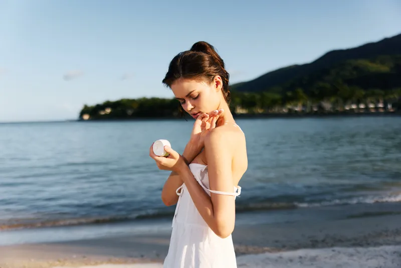 Young woman applying sunscreen lotion on her shoulder at the beach during summer, with calm sea and mountains in the background.
