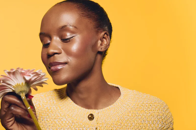 Portrait of a happy African woman with dark skin holding a flower, wearing a yellow sweater on yellow background. Concept of natural beauty, wellness, and floral scent enjoyment.