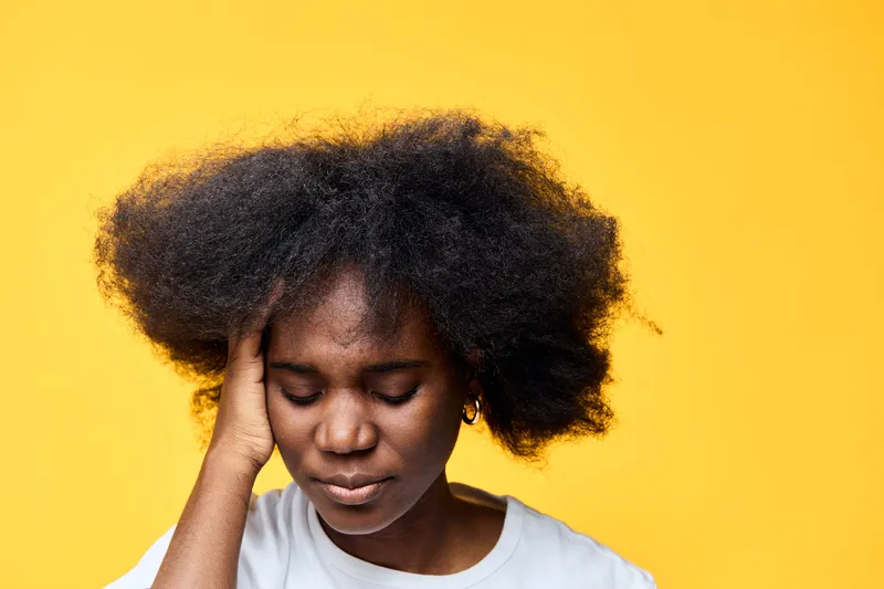 Young African woman with curly hair experiencing stress, against a vibrant yellow background, conveying feelings of anxiety and concern