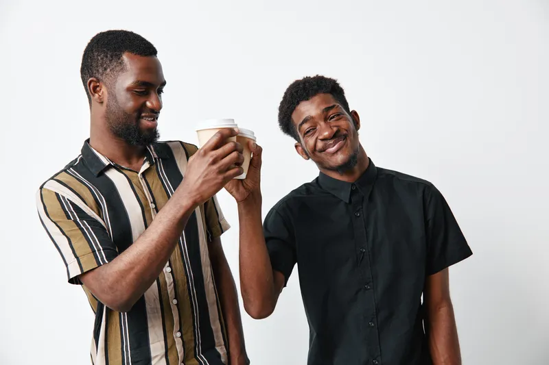 Two young black men with dark skin smiling and holding coffee cups together in casual clothing on white background. Friendship, happiness and relaxed mood in studio.