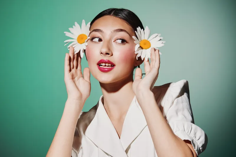 Young woman with glowing skin and vibrant red lips poses playfully holding daisies near her ears. Bright studio lighting emphasizes fresh beauty and creative fashion concept.
