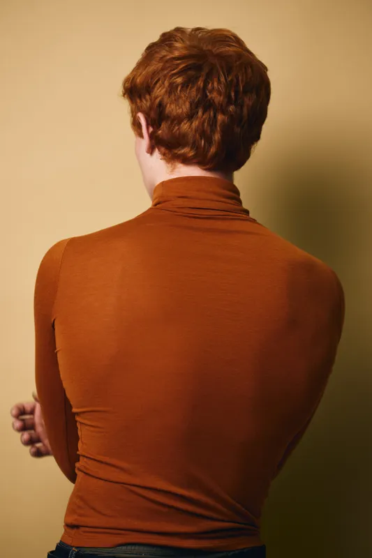Person with red hair facing away in brown turtleneck sweater against beige background. Casual style with folded arms and short hairstyle showing back view indoors.